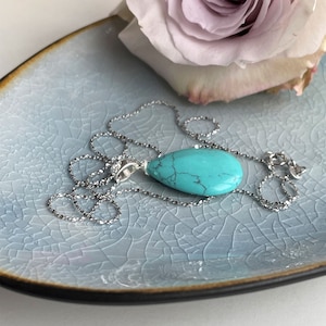 May include: A turquoise teardrop pendant necklace with a silver chain rests on a light blue, crackle-glazed ceramic dish. A soft pink rose is in the background, adding a touch of color to the scene. The necklace is a simple, elegant piece.