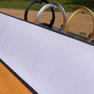 K&ouml;nnte beinhalten: Ein langes, rechteckiges Display-Board mit einer wei&szlig;en Stoffoberfl&auml;che und einem dunklen Rahmen. Drei halbkreisf&ouml;rmige Metallst&auml;nder in den Farben Schwarz, Silber und Gold sind entlang der Oberkante angebracht und dienen als stabile Basis.
