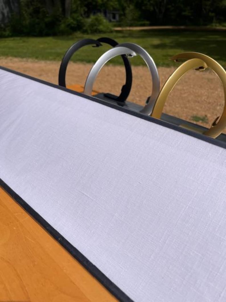 K&ouml;nnte beinhalten: Ein langes, rechteckiges Display-Board mit einer wei&szlig;en Stoffoberfl&auml;che und einem dunklen Rahmen. Drei halbkreisf&ouml;rmige Metallst&auml;nder in den Farben Schwarz, Silber und Gold sind entlang der Oberkante angebracht und dienen als stabile Basis.