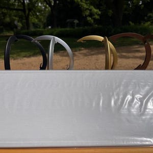 K&ouml;nnte beinhalten: Ein Set aus vier geschwungenen Metallhaken in den Farben Schwarz, Silber, Gold und Bronze, die in einer Reihe hinter einer rechteckigen, hellgrauen, strukturierten Oberfl&auml;che mit schwacher, gepr&auml;gter Schrift auf der Vorderseite stehen.