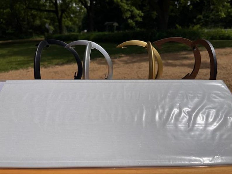K&ouml;nnte beinhalten: Ein Set aus vier geschwungenen Metallhaken in den Farben Schwarz, Silber, Gold und Bronze, die in einer Reihe hinter einer rechteckigen, hellgrauen, strukturierten Oberfl&auml;che mit schwacher, gepr&auml;gter Schrift auf der Vorderseite stehen.