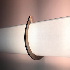 May include: A wall-mounted light fixture featuring a long, horizontal, cylindrical white fabric shade. The shade is held in place by a curved, dark brown wooden bracket that wraps around the center, creating a sleek and functional lighting design.