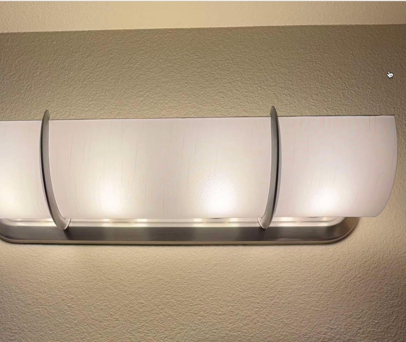 K&ouml;nnte beinhalten: Eine horizontale Wandleuchte mit einem langen, geschwungenen, mattwei&szlig;en Glasschirm. Der Schirm wird von zwei Halterungen aus geb&uuml;rstetem Nickel gehalten, die sich um die Vorderseite legen und an einer passenden Grundplatte befestigt sind.