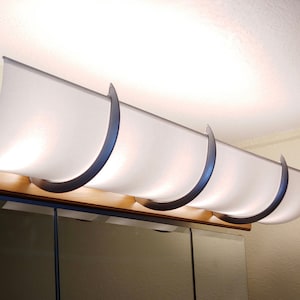 May include: A horizontal wall-mounted light fixture featuring a long, curved, frosted white glass shade. The shade is held in place by three dark gray, semi-circular metal brackets attached to a light-toned wooden base plate mounted above a mirror.