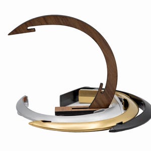 K&ouml;nnte beinhalten: Ein geschwungener Holzst&auml;nder mit einer spitzen Oberseite, platziert hinter einem Stapel aus drei kreisf&ouml;rmigen Ringen mit metallischem Finish in Silber, Gold und Schwarz. Die Ringe sind ineinander verschachtelt und der Holzst&auml;nder hat eine Kerbe.