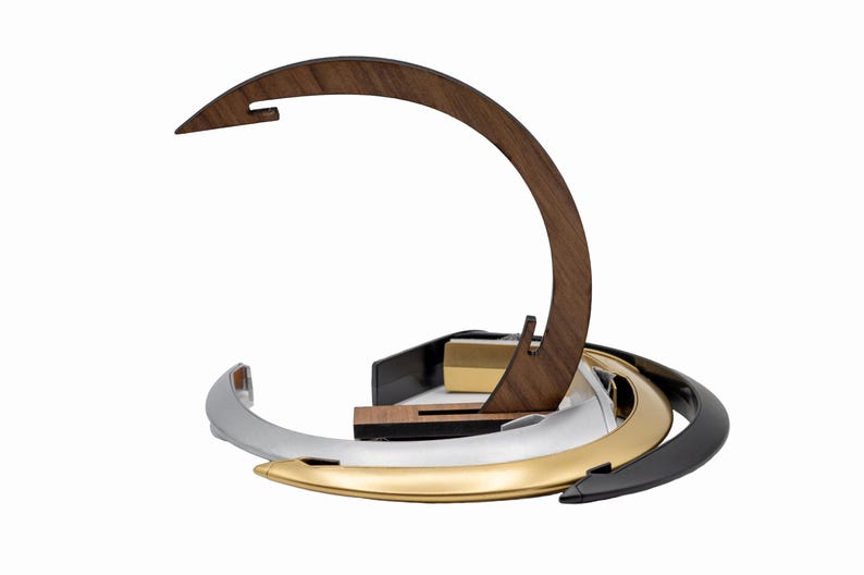 K&ouml;nnte beinhalten: Ein geschwungener Holzst&auml;nder mit einer spitzen Oberseite, platziert hinter einem Stapel aus drei kreisf&ouml;rmigen Ringen mit metallischem Finish in Silber, Gold und Schwarz. Die Ringe sind ineinander verschachtelt und der Holzst&auml;nder hat eine Kerbe.