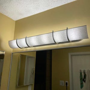 May include: A rectangular, gray fabric bathroom light fixture with a curved design. The light fixture is mounted above a mirror and has a silver metal frame. The light is on, illuminating the room.