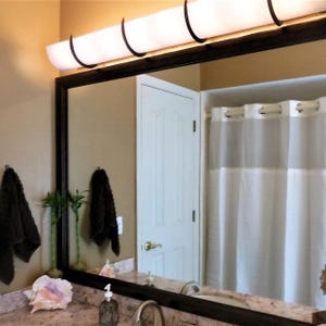 May include: A bathroom vanity with a large mirror, a white shower curtain, and a light fixture with multiple curved bulbs. The mirror has a dark wood frame and reflects a white door and a brown wall.