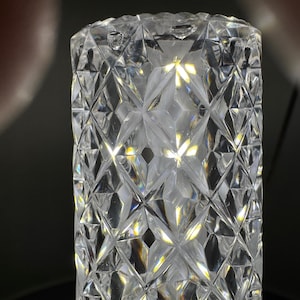 May include: A clear, cylindrical glass object with a diamond-cut pattern. The facets catch and reflect light, creating a sparkling effect. The object is likely a decorative vase or container.