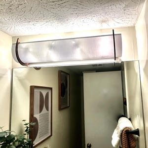 May include: A rectangular, frosted glass bathroom light fixture with curved, dark brown metal accents. The light is mounted above a large mirror, reflecting the fixture and surrounding decor.
