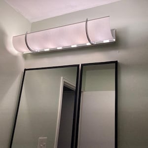 May include: A horizontal wall-mounted light fixture featuring a curved, off-white fabric shade held in place by two silver-toned metal brackets. The fixture is positioned above two rectangular mirrors with thin black frames on a light-colored wall.