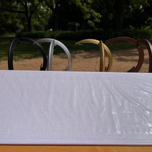 K&ouml;nnte beinhalten: Vier geschwungene Metallst&auml;nder in einer Reihe angeordnet, mit matten Oberfl&auml;chen in Schwarz, Silber, Gold und Dunkelbraun. Jeder St&auml;nder verf&uuml;gt &uuml;ber einen abgerundeten oberen Haken und einen stabilen Fu&szlig;, hinter einer rechteckigen wei&szlig;en Platte.