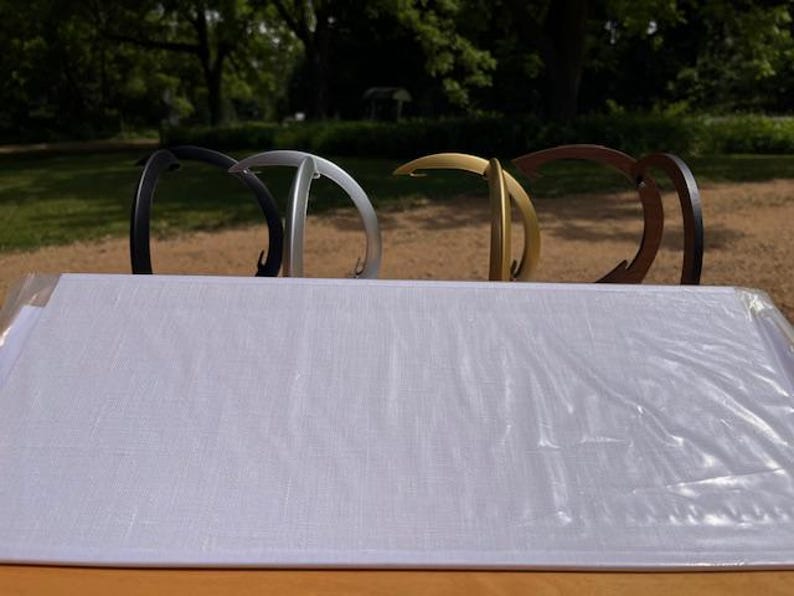 K&ouml;nnte beinhalten: Vier geschwungene Metallst&auml;nder in einer Reihe angeordnet, mit matten Oberfl&auml;chen in Schwarz, Silber, Gold und Dunkelbraun. Jeder St&auml;nder verf&uuml;gt &uuml;ber einen abgerundeten oberen Haken und einen stabilen Fu&szlig;, hinter einer rechteckigen wei&szlig;en Platte.