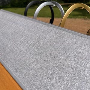 K&ouml;nnte beinhalten: Ein rechteckiges Display-Board mit einer strukturierten hellgrauen Stoffoberfl&auml;che und einem silbernen Metallrahmen. Hinter dem Board sind drei bogenf&ouml;rmige Metallgriffe in den Farben Schwarz, Silber und Gold in einer Reihe angeordnet.