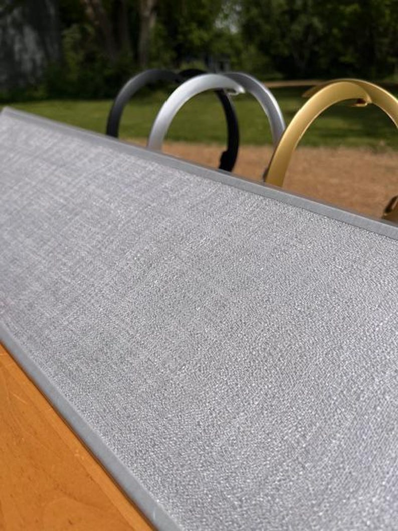 K&ouml;nnte beinhalten: Ein rechteckiges Display-Board mit einer strukturierten hellgrauen Stoffoberfl&auml;che und einem silbernen Metallrahmen. Hinter dem Board sind drei bogenf&ouml;rmige Metallgriffe in den Farben Schwarz, Silber und Gold in einer Reihe angeordnet.