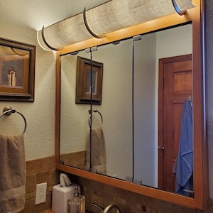 May include: A rectangular medicine cabinet with a light wood frame and three mirrored doors. Above the mirror is a horizontal light fixture with a textured, beige fabric shade held by dark metal brackets, providing warm illumination.