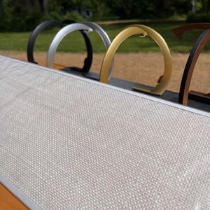 K&ouml;nnte beinhalten: Ein rechteckiges Board mit einer hellgrauen, strukturierten Stoffoberfl&auml;che und einem silberfarbenen Metallrahmen. Hinter dem Board ist eine Reihe von kreisf&ouml;rmigen Metallst&auml;ndern in Schwarz, Silber, Gold und Bronze aufgereiht.