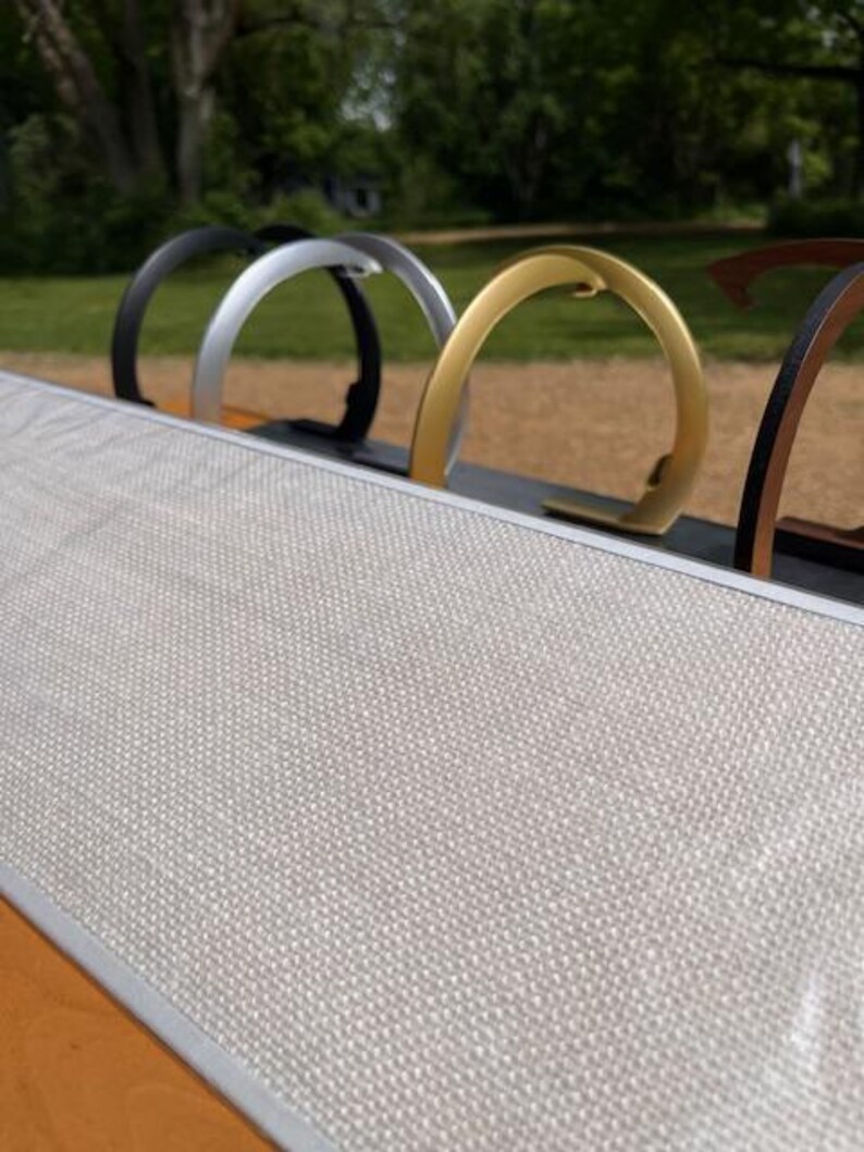 K&ouml;nnte beinhalten: Ein rechteckiges Board mit einer hellgrauen, strukturierten Stoffoberfl&auml;che und einem silberfarbenen Metallrahmen. Hinter dem Board ist eine Reihe von kreisf&ouml;rmigen Metallst&auml;ndern in Schwarz, Silber, Gold und Bronze aufgereiht.