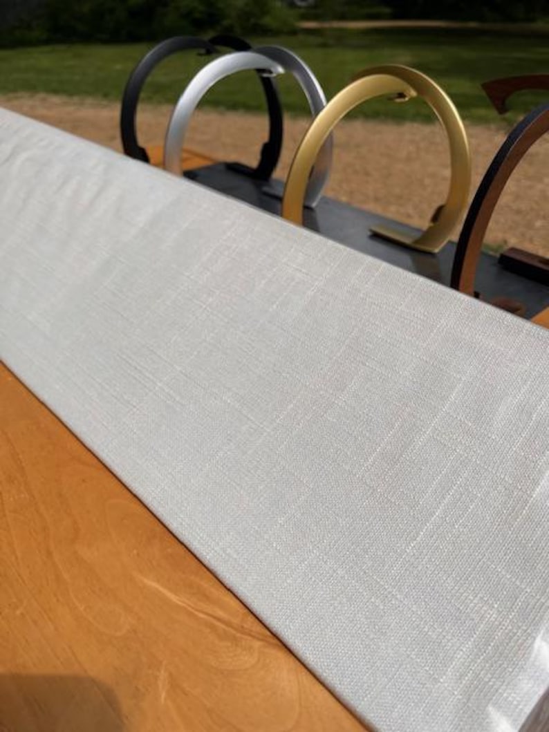 K&ouml;nnte beinhalten: Ein rechteckiges, hellgraues, mit strukturiertem Stoff bezogenes Display-Board, das schr&auml;g aufgestellt ist. Dahinter sind mehrere bogenf&ouml;rmige, halbkreisf&ouml;rmige Metallst&auml;nder in den Farben Schwarz, Silber, Gold und Dunkelbraun in einer Reihe angeordnet.