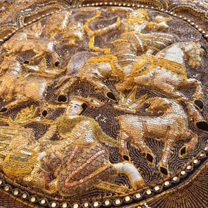 Könnte beinhalten: Nahaufnahme eines kunstvollen Textils mit goldenen Pailletten und Stickereien. Das Design zeigt Figuren auf Pferden, Bogenschützen und andere detaillierte Elemente. Der Hintergrund ist dunkelbraun mit wirbelnden Mustern.