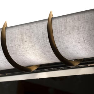 May include: Close-up of a window treatment featuring a light gray fabric shade. The shade is held in place by curved, bronze-colored metal brackets. The fabric has a textured appearance, and the design is simple and elegant.