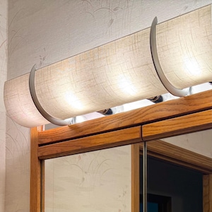 May include: A bathroom vanity light fixture with a beige fabric shade and three curved metal supports. The fixture is mounted above a wooden cabinet with a mirrored door. The light is turned on, illuminating the space.