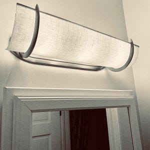 May include: A rectangular wall-mounted light fixture with a fabric shade and a metal frame. The shade is a light beige color, and the frame has a silver finish. The fixture is mounted above a mirror.