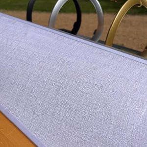 K&ouml;nnte beinhalten: Eine rechteckige Anzeigetafel mit einer hellgrauen, strukturierten Stoffoberfl&auml;che und einem d&uuml;nnen silbernen Rahmen. Hinter der Tafel sind drei bogenf&ouml;rmige Metallst&auml;nder in Schwarz, Silber und Gold in einer Reihe vor einem unscharfen Hintergrund zu sehen.