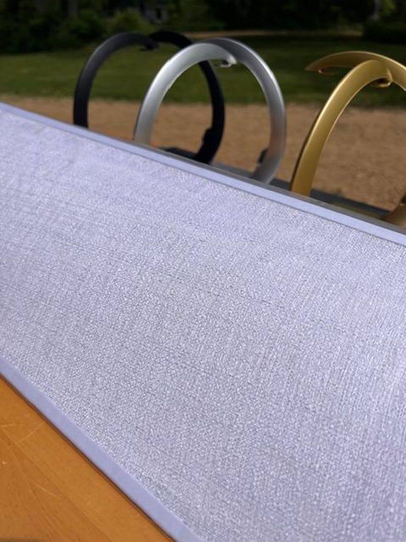 K&ouml;nnte beinhalten: Eine rechteckige Anzeigetafel mit einer hellgrauen, strukturierten Stoffoberfl&auml;che und einem d&uuml;nnen silbernen Rahmen. Hinter der Tafel sind drei bogenf&ouml;rmige Metallst&auml;nder in Schwarz, Silber und Gold in einer Reihe vor einem unscharfen Hintergrund zu sehen.