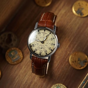 May include: A silver wristwatch with a white face and black hands. The watch has a brown leather band and a circular face with black numbers and markings. The watch is on a wooden surface.