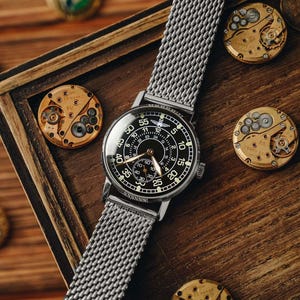 May include: A silver wristwatch with a black face and mesh band rests on a wooden surface. The watch face displays white numerals and hands. Several watch movement components and bottle caps are scattered around the watch.