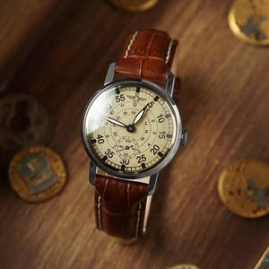 Puede incluir: Un reloj de estilo vintage con esfera crema y n&uacute;meros negros. El reloj tiene una caja de acero inoxidable y una correa de cuero marr&oacute;n. La esfera muestra m&uacute;ltiples esferas y agujas, sobre una superficie de madera.