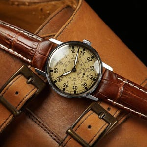 Puede incluir: Un reloj de pulsera vintage con esfera clara y n&uacute;meros negros, con una correa de cuero marr&oacute;n con estampado de cocodrilo. El reloj se encuentra sobre un zapato de cuero marr&oacute;n, con hebillas met&aacute;licas.
