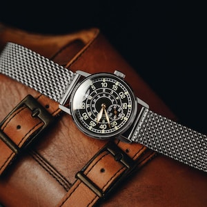 May include: A wristwatch with a round black dial featuring white Arabic numerals for minutes and a smaller inner circle for hours. The watch has gold-toned hands and a silver-toned stainless steel mesh strap with a polished finish and a circular case.