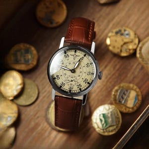 Puede incluir: Un reloj de pulsera de estilo vintage con una correa de cuero marr&oacute;n y una esfera color crema. El reloj tiene una caja plateada y una peque&ntilde;a subesfera. El reloj est&aacute; rodeado de monedas antiguas sobre una superficie de madera.