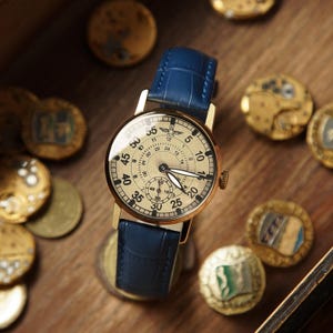 May include: A gold-toned wristwatch with a cream-coloured face and a blue leather strap. The watch face displays numbers and smaller dials. The watch is surrounded by gold-coloured coins on a wooden surface.