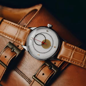 May include: A silver wristwatch with a white face, black and gold accents, and a red second hand rests on a brown leather strap. The watch is set on a pair of brown leather shoes with buckle closures, showcasing a classic style.