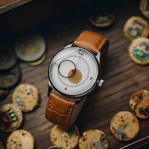 May include: A silver wristwatch with a textured white face, black and red accents, and a brown leather band. The watch is surrounded by gold-colored coins, set in a wooden box.