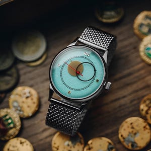 Op de afbeelding: Een zilveren polshorloge met een mesh band en een lichtblauwe wijzerplaat. De wijzerplaat heeft een goudkleurige cirkel, een zwarte cirkel en een rode wijzer. Het horloge ligt op een houten oppervlak met munten.