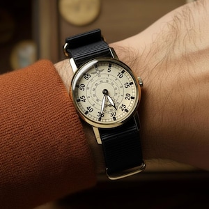 May include: A gold-toned wristwatch with a black nylon strap. The watch face is cream-colored with black numerals and hands. The watch is on a person's wrist, showing the time. The watch has a classic, vintage aesthetic.