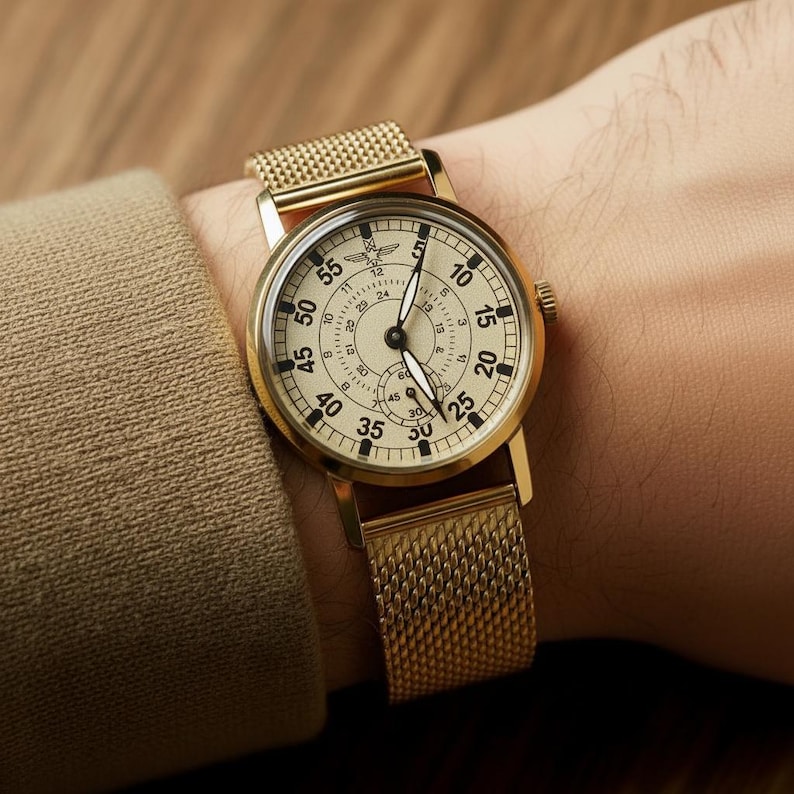 Peut inclure: Montre-bracelet dor&eacute;e avec un bracelet en maille. Le cadran de la montre est de couleur cr&egrave;me avec des chiffres et des aiguilles noirs. La montre est port&eacute;e au poignet, et l'esth&eacute;tique g&eacute;n&eacute;rale est vintage et classique.