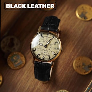 May include: A gold-coloured wristwatch with a black leather strap. The watch face is cream with black numerals and hands. The text "BLACK LEATHER" is at the top. The watch is on a wooden surface.