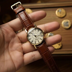 May include: A gold-toned wristwatch with a cream-colored face and black numerals. The watch has a brown leather band with a crocodile pattern. The watch face displays multiple dials and hands, and the watch is held in a hand.