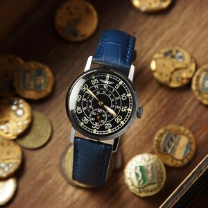 May include: A black-faced wristwatch with a blue leather band. The watch has a silver case and a detailed dial with numbers and hands. The watch is surrounded by gold coins on a wooden surface.