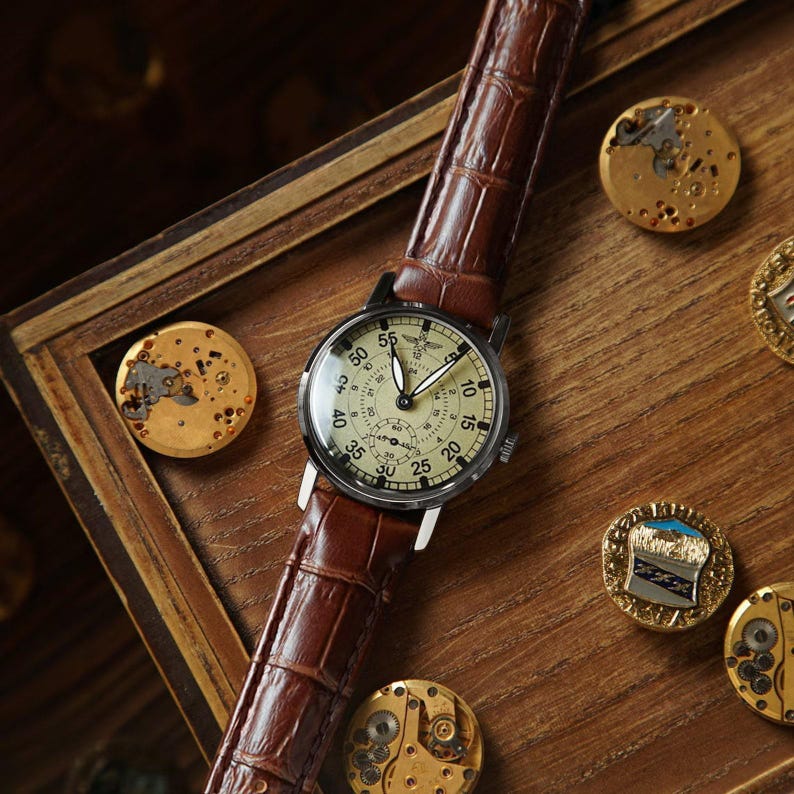 Puede incluir: Un reloj de pulsera de estilo vintage con esfera crema y n&uacute;meros negros, con una correa de cuero marr&oacute;n con estampado de cocodrilo. El reloj est&aacute; sobre una superficie de madera, rodeado de componentes de reloj y elementos decorativos.