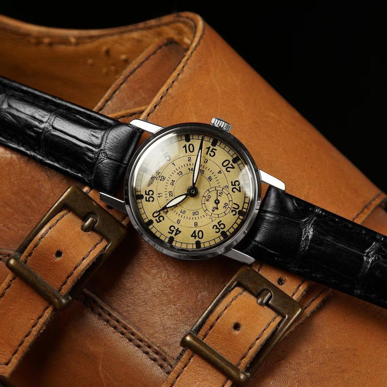 May include: A vintage wristwatch with a cream-colored face and black leather band rests on a brown leather shoe. The watch has a silver case and features multiple dials and numbers. The shoe has two buckles.