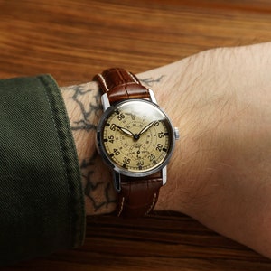 May include: A silver wristwatch with a brown leather band. The watch face is cream colored with black markings and a 24-hour dial. The watch is on a person's wrist.