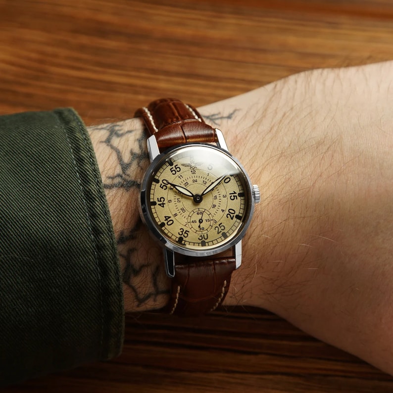 May include: A silver wristwatch with a brown leather band. The watch face is cream colored with black markings and a 24-hour dial. The watch is on a person's wrist.