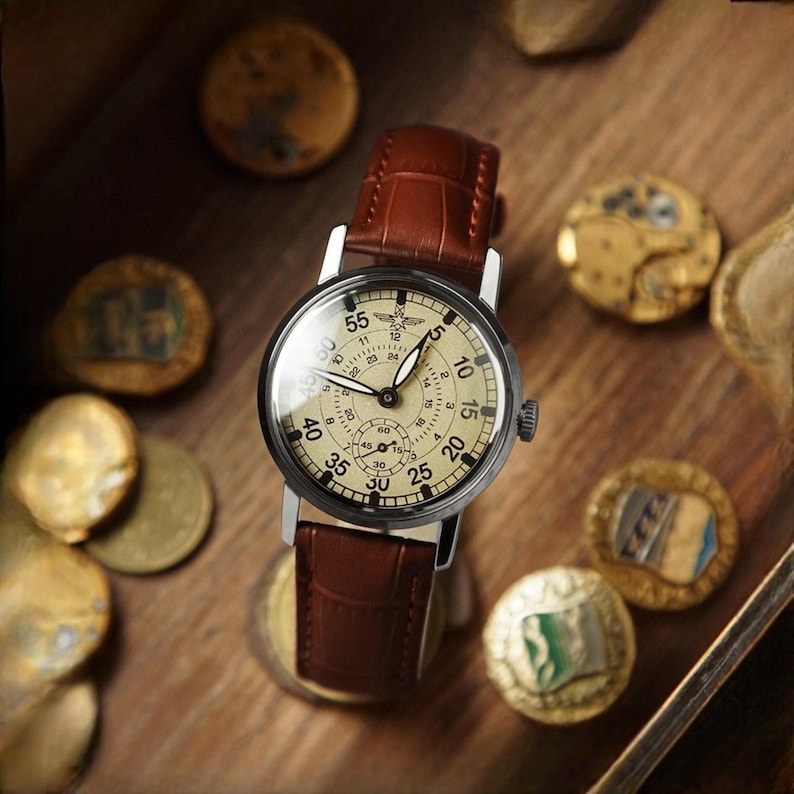 Puede incluir: Un reloj de pulsera de estilo vintage con una correa de cuero marr&oacute;n y una esfera color crema. El reloj tiene una caja plateada y una peque&ntilde;a subesfera. El reloj est&aacute; rodeado de monedas antiguas sobre una superficie de madera.