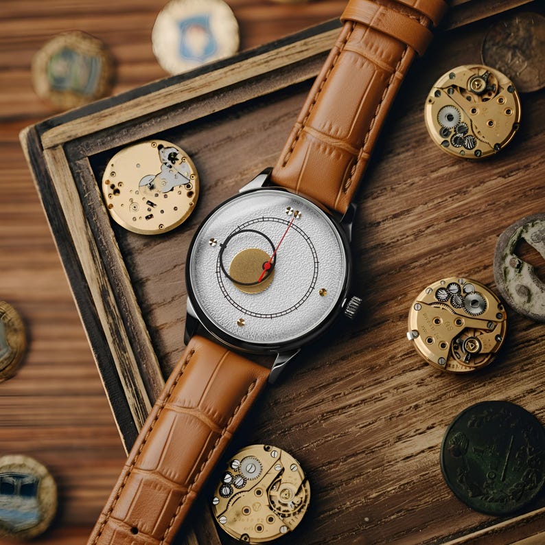 May include: A wristwatch with a textured white dial, a black circular sub-dial, and a thin red hand. The watch features a tan leather strap with a crocodile-embossed pattern. Several circular watch movement components are scattered nearby.